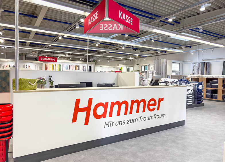 Hammer Fachmarkt: Das Bild zeigt den Kassenbereich eines Hammer-Fachmarktes mit einem großen Schild "Hammer - Mit uns zum TraumRaum" sowie mehreren roten Kassenschildern und Regalen mit Produkten im Hintergrund.